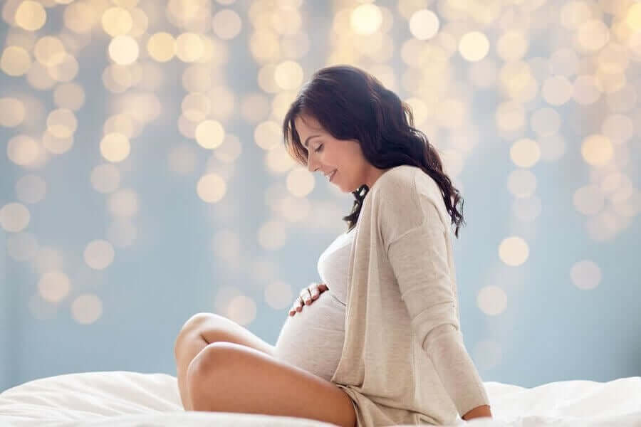 Gravid kvinde sidder i skrædderstilling og holder på sin mave med et smil.