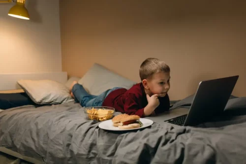 Dreng med computer og burger i seng repræsenterer, hvordan nogle spiser foran skærmene